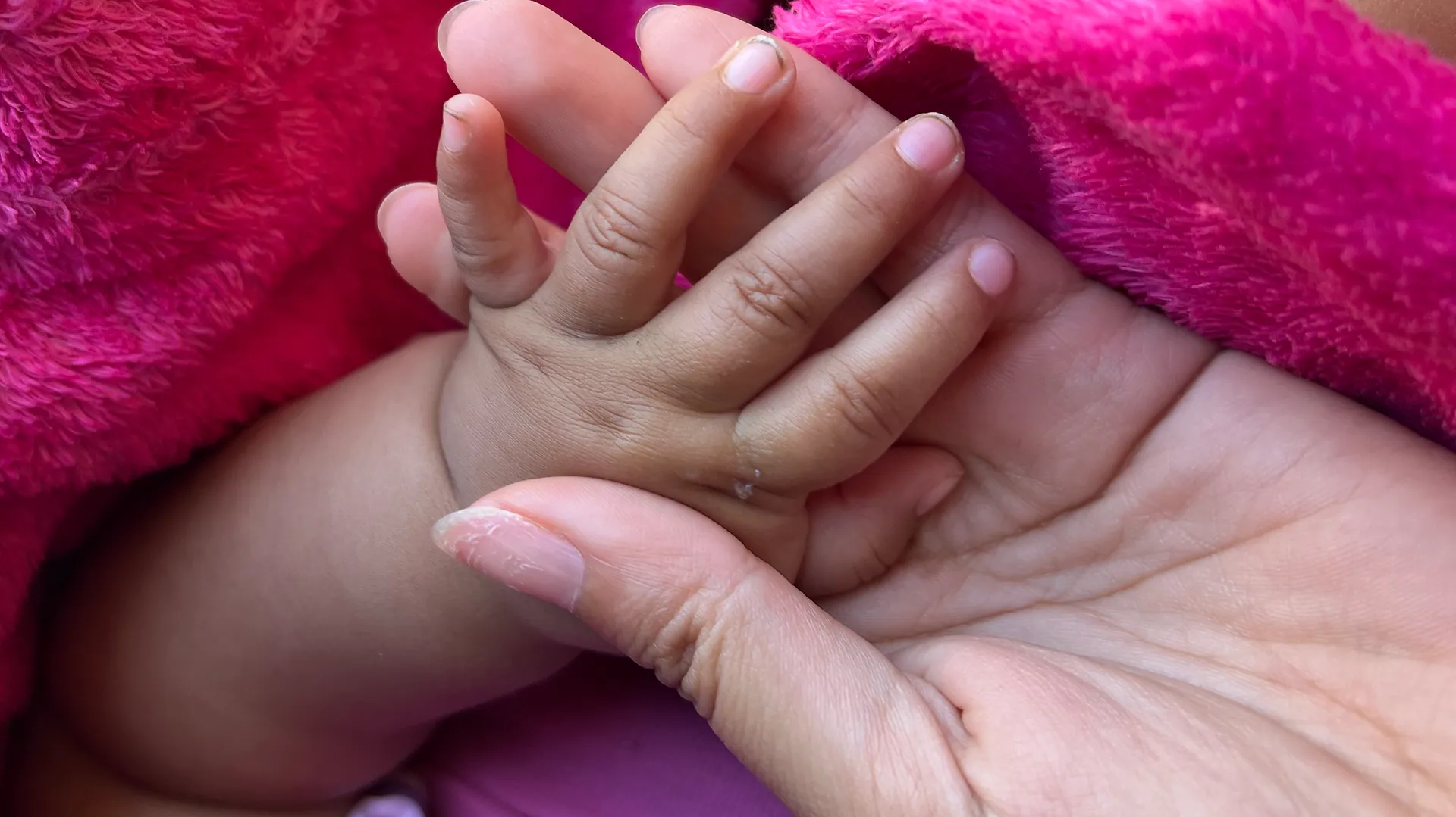A baby's small hand resting in a woman's open palm, wrapped in a soft pink blanket