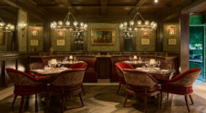 Main dining room with wood paneling, chandeliers, curved leather banquette and red club chairs