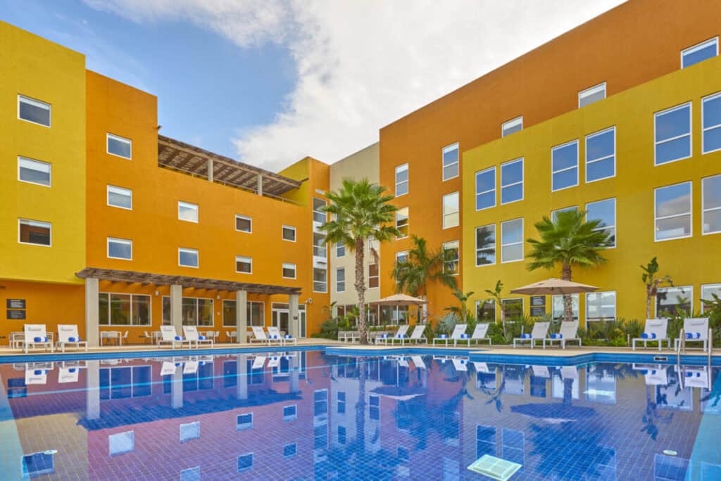 City Express Plus & Suites Cabo San Lucas — orange and yellow hotel block behind a rectangular pool with loungers