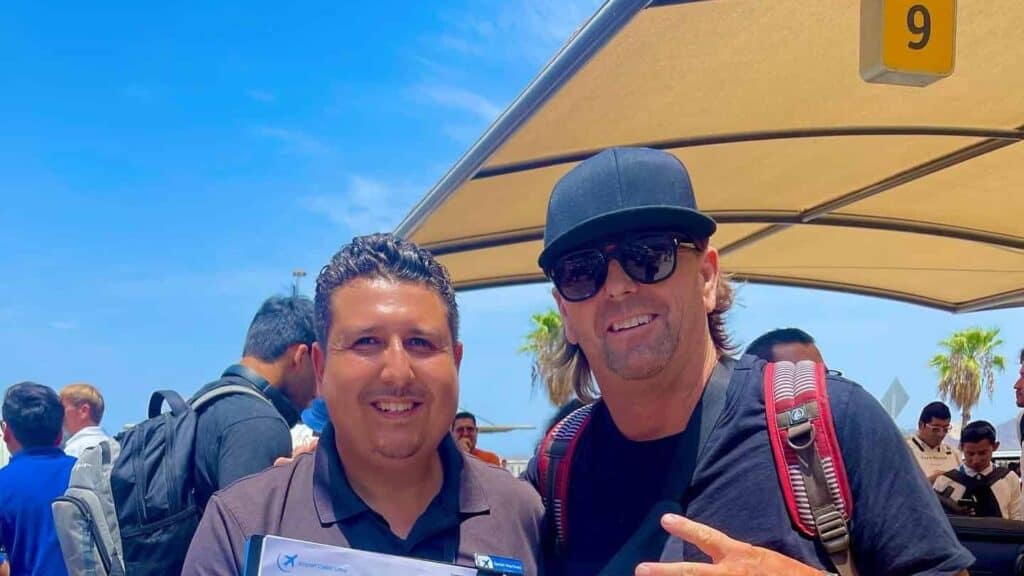 A driver holding an Airport Cabo Limo clipboard poses with a guest wearing a backpack under shade canopy number 9