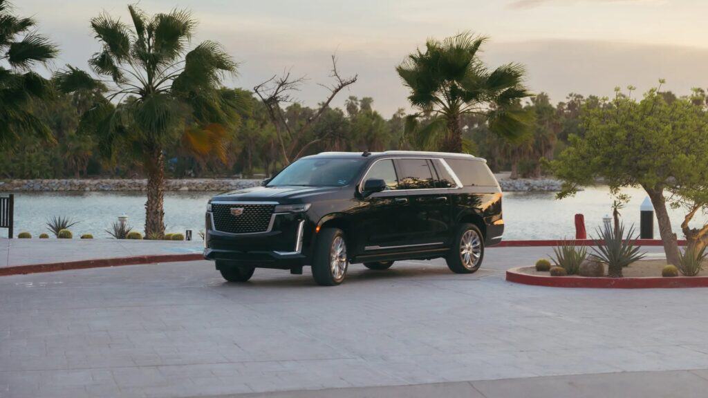 A black Cadillac Escalade parked by palm trees and calm water at dusk