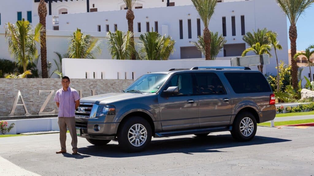 A driver standing beside a gray Ford Expedition parked in front of a white resort building with palm trees