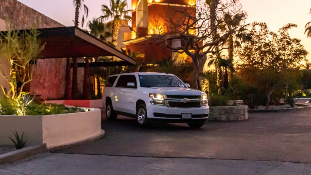 A white Cabo's Routes Suburban with its headlights on at dusk, parked by a lit restaurant entrance with palm trees behind