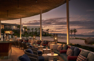 Sunset view of the lounge and bar seating area at Casa de Brasa, with the lit bar, cushioned chairs and the Baja coastline at dusk
