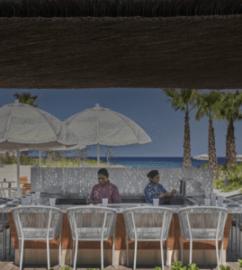 Beachfront bar counter at Casa de Brasa with staff at work, white woven umbrellas, palms and the Sea of Cortez in the background
