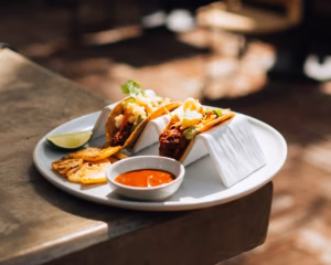 Three tacos on a white plate with grilled pineapple, lime, and red salsa