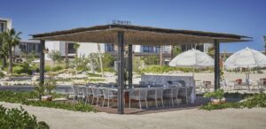 El Puesto beachside palapa with the venue name on the roof, sand-floor dining tables and shade umbrellas at the Marina Village