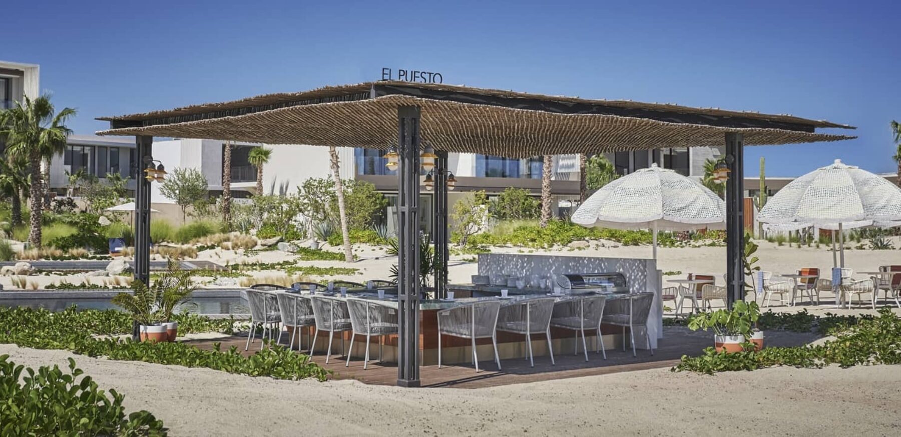 El Puesto beachside palapa with the venue name on the roof, sand-floor dining tables and shade umbrellas at the Marina Village