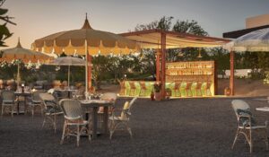 Open-air orchard dining room at dusk: linen umbrellas, rattan chairs and the glowing copper-framed bar (official Costa Palmas media, 1800x1050)