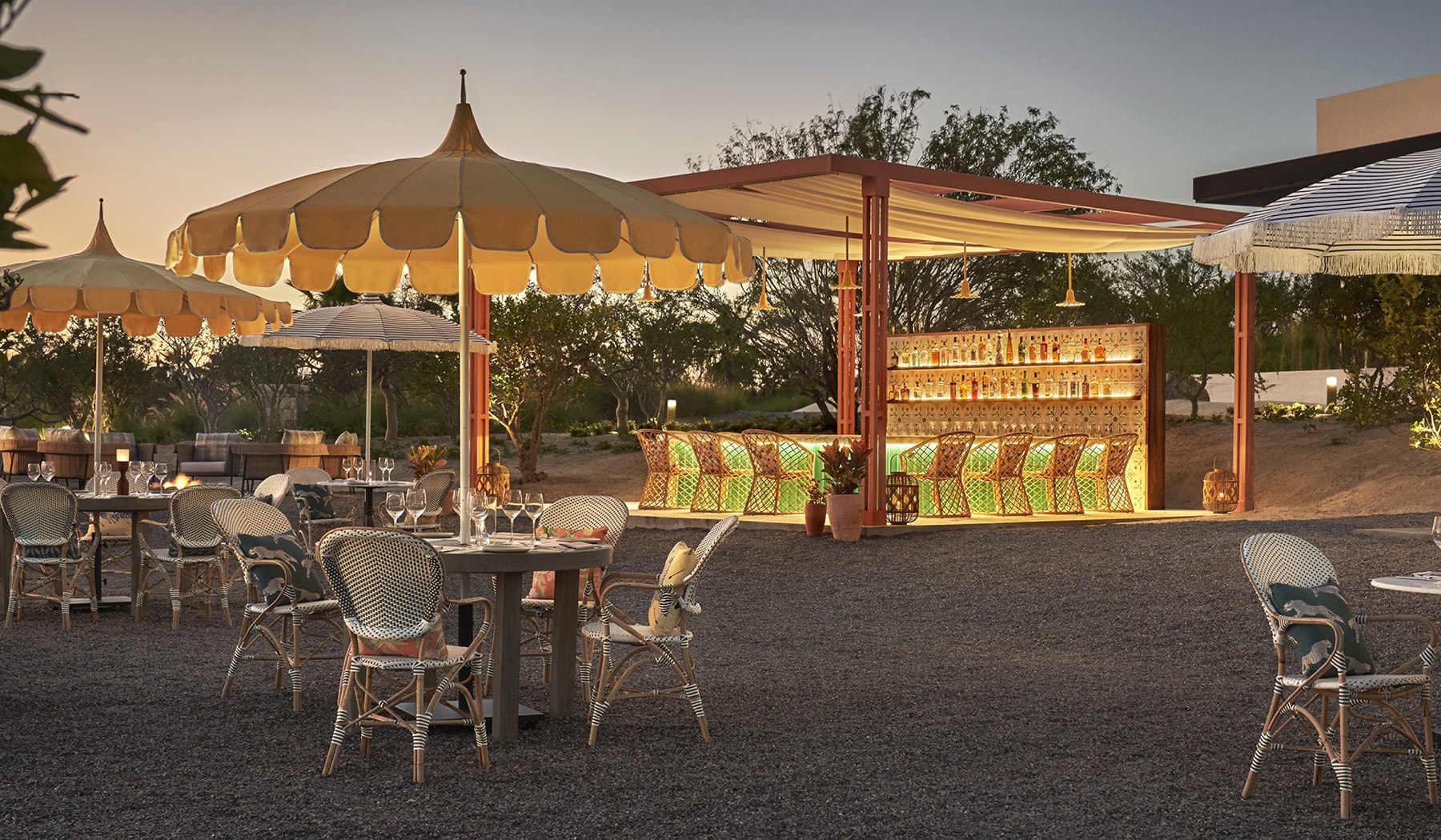 Open-air orchard dining room at dusk: linen umbrellas, rattan chairs and the glowing copper-framed bar (official Costa Palmas media, 1800x1050)