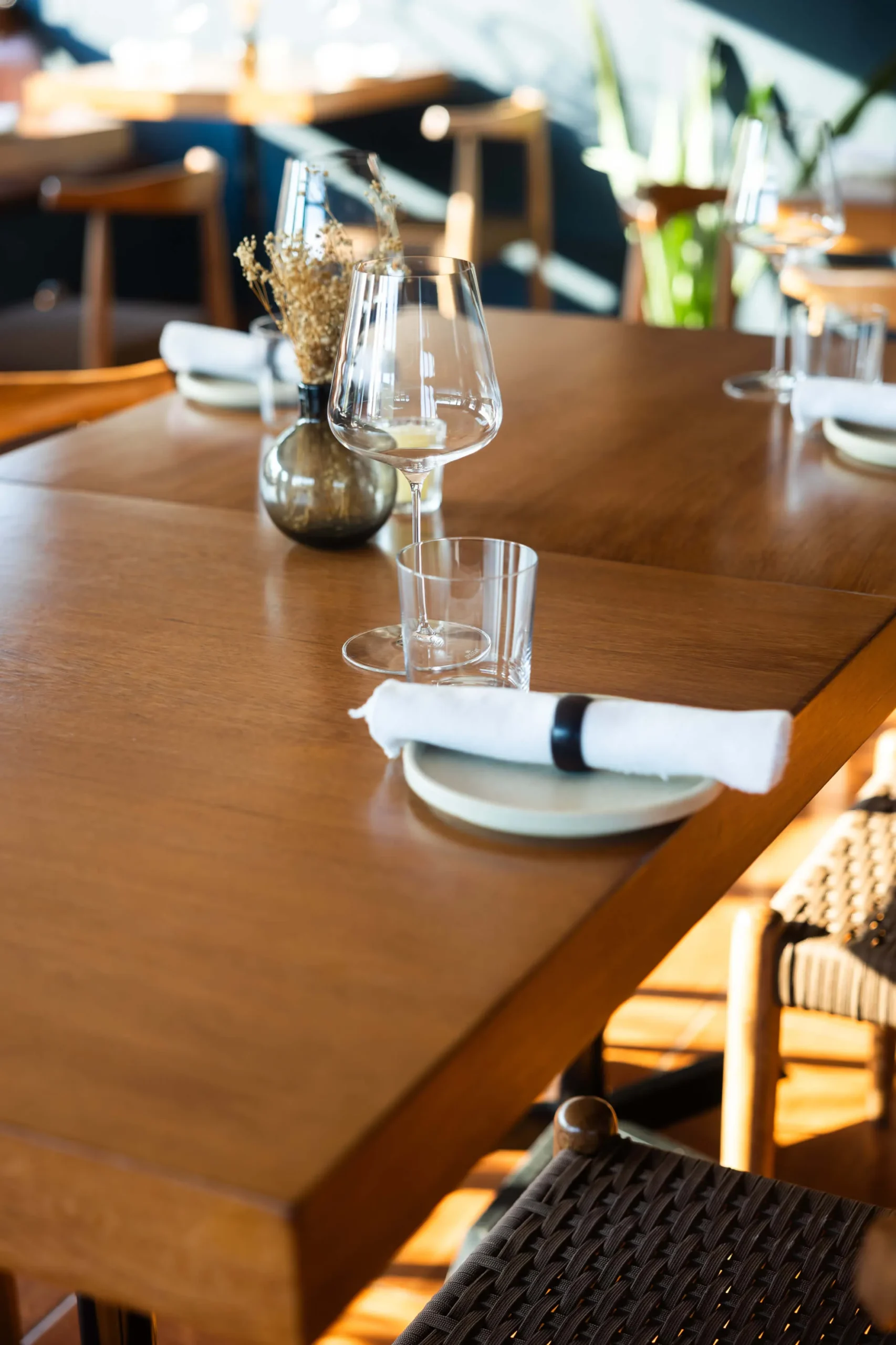 Official site hero: warm timber dining table set with wine glass, dried-flower vase and linen in a sunlit dining room
