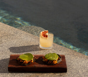 Citrus cocktail and avocado-topped tostadas plated on a wood board at the pool edge