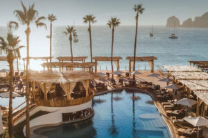 Beach club pool, palapas and palms with Land's End rock formations and yachts beyond