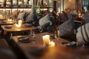 Candlelit dinner tables with cushions and wine glasses in front of the lit bar