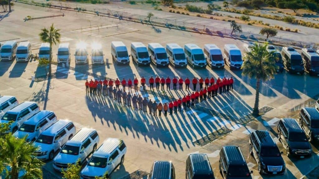 Aerial view of Transcabo staff in red and grey uniforms standing in a heart shape in a parking lot, surrounded by rows of white vans, white SUVs and black Cadillac Escalades