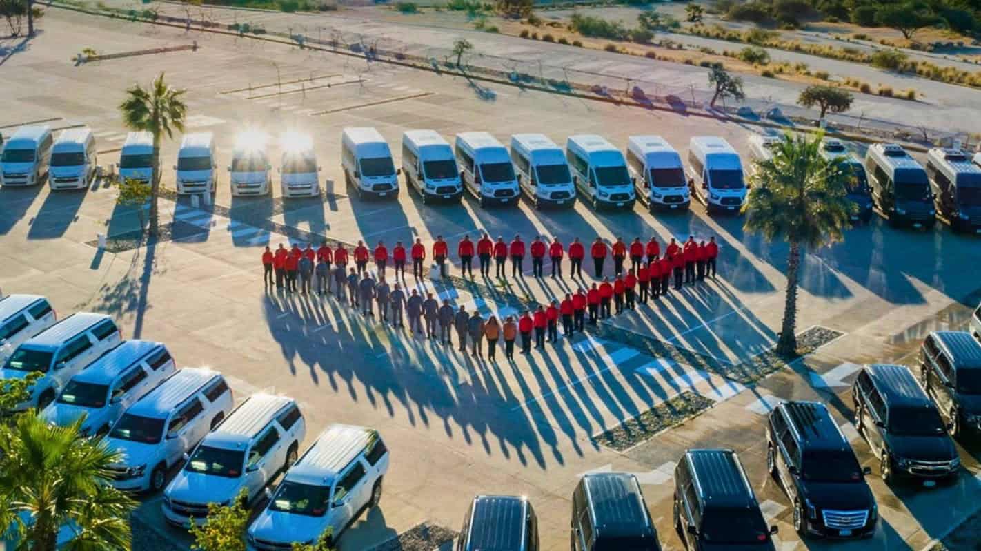 Aerial view of Transcabo staff in red and grey uniforms standing in a heart shape in a parking lot, surrounded by rows of white vans, white SUVs and black Cadillac Escalades