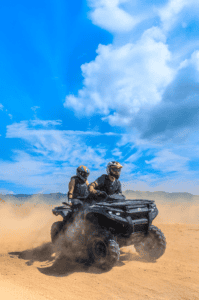 Two helmeted guests riding an automatic ATV across sandy terrain