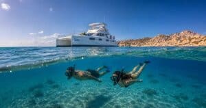 Two snorkelers swimming below a Cabo Adventures catamaran in clear water