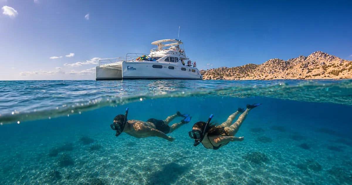 Two snorkelers swimming below a catamaran in clear Los Cabos water