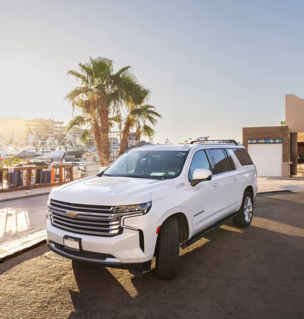 A white Chevrolet Suburban on a sand road in Los Cabos, palms and low buildings behind.