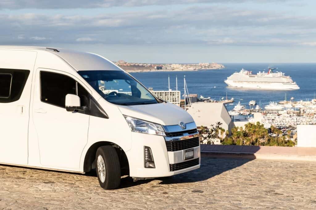 A white passenger van parked above the Cabo San Lucas marina, a cruise ship anchored offshore.