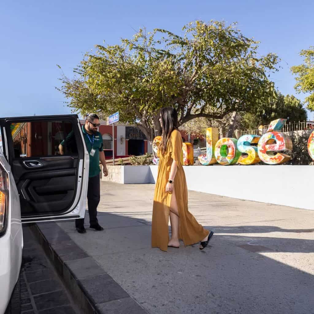 A driver in uniform holding the door of a white SUV as a guest walks up in San Jose del Cabo.