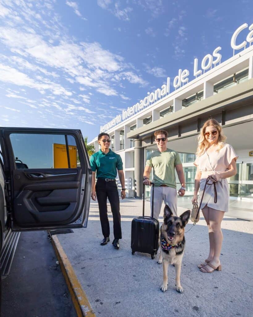 Three travellers and a dog with luggage outside the Los Cabos airport terminal, next to an open van door.