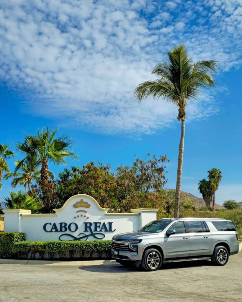 A white SUV at the Cabo Real entrance sign, palms and blue sky behind.