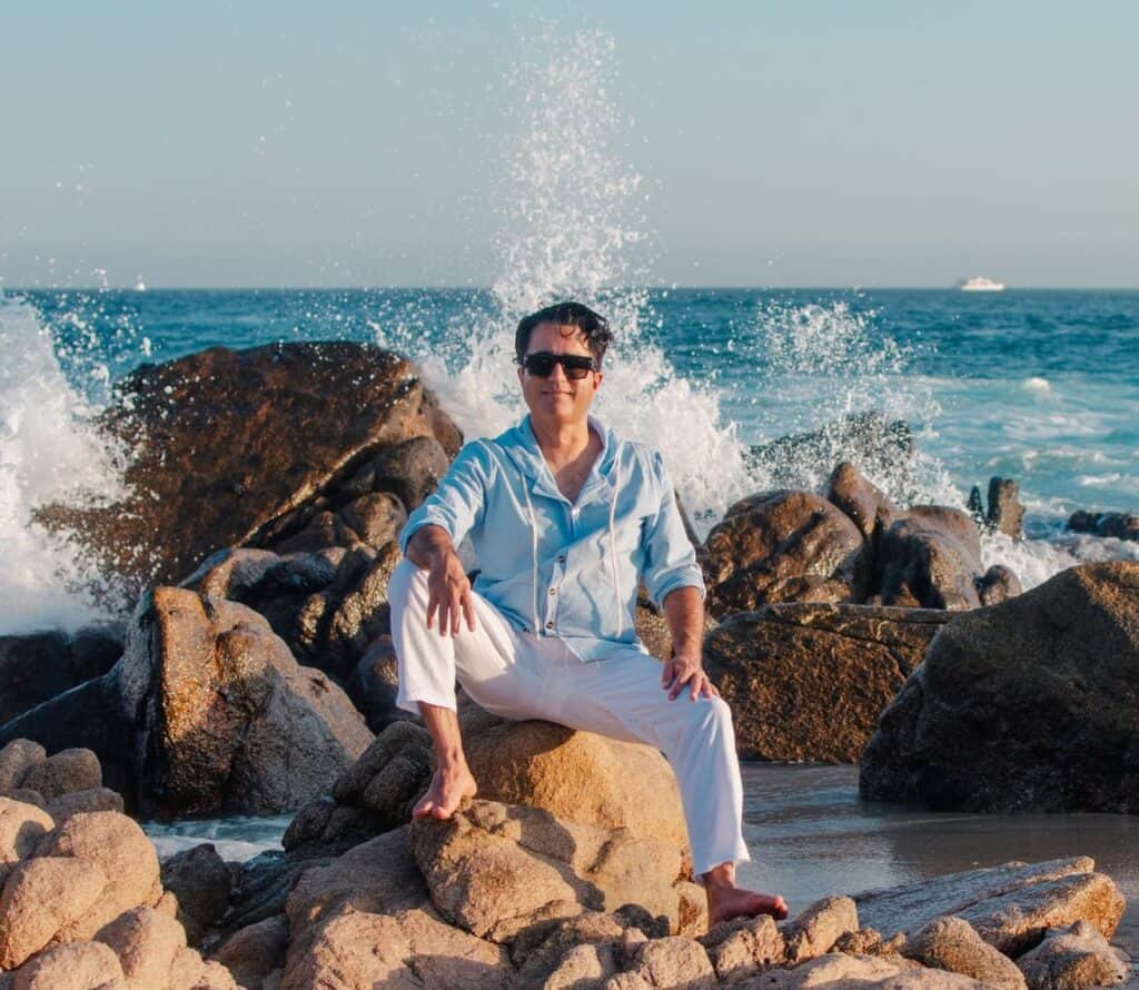 Man sitting on rocks at Cabo San Lucas as a wave breaks behind him