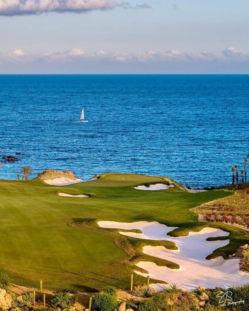 A green ringed by bunkers with turquoise water and a sailboat beyond