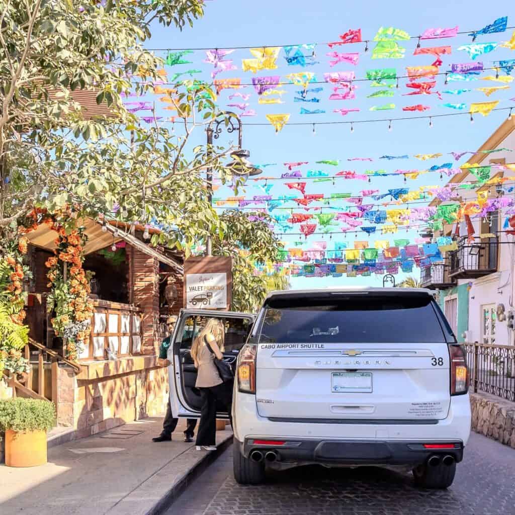 A white shuttle parked on a San Jose del Cabo street hung with papel picado while a passenger steps out.