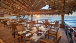 The palapa dining room at 7 Seas Seafood Grille with tables set and the sun low over the sea at Acapulquito Beach