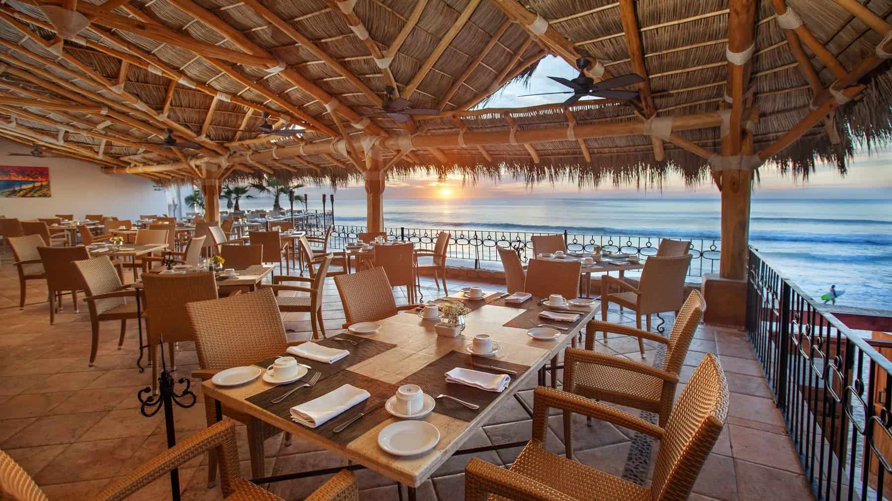 The palapa dining room at 7 Seas Seafood Grille with tables set and the sun low over the sea at Acapulquito Beach