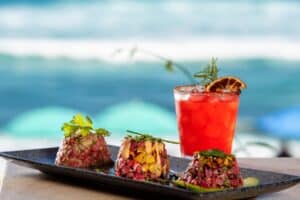 Three tuna tartare towers beside a red cocktail with a dried orange slice, with the surf behind, at 7 Seas