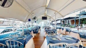 The covered deck at Baja Lobster Co. with tables set in blue linen and boats moored along the Cabo San Lucas marina