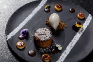 A small sugar-dusted cake with a quenelle of ice cream on crumble and caramelized rounds on a dark slate plate at Café des Artistes Los Cabos
