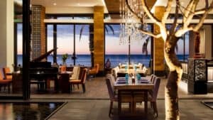 The dining room at Café des Artistes Los Cabos at dusk, with a grand piano, set tables and the Sea of Cortez through the glass walls