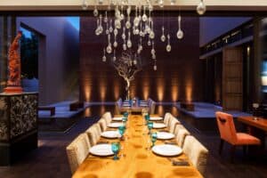 A long wooden table set with blue glassware under hanging glass-drop lamps at Café des Artistes Los Cabos