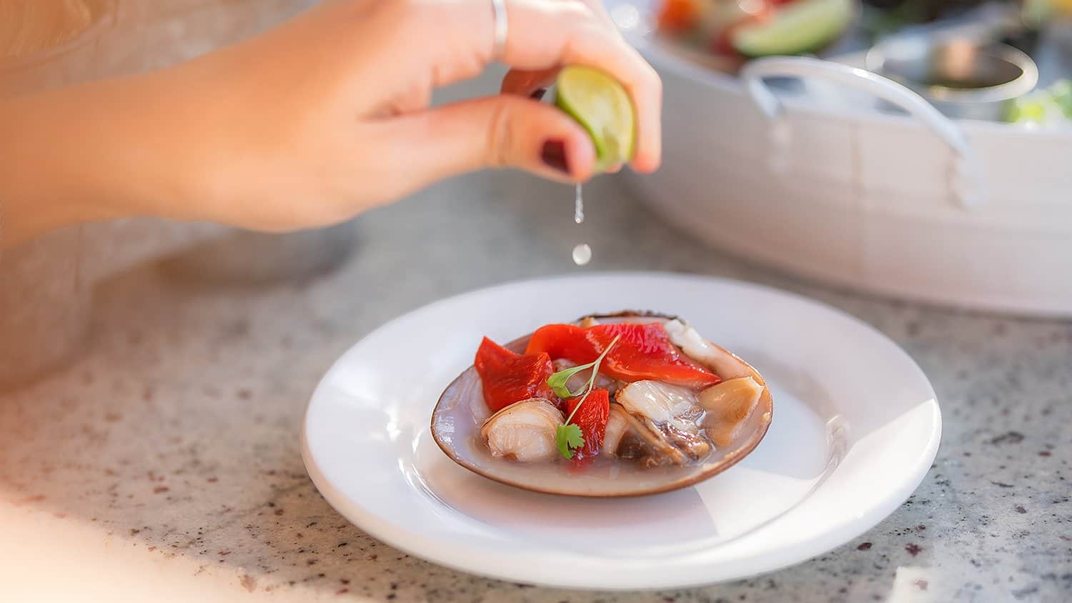 A squeeze of lime over a raw clam on the half shell, with a platter of shellfish on ice behind, at Casamarte in La Paz