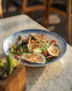 Grilled oysters with a golden crust on a salad of greens, cherry tomatoes and red onion in a blue-rimmed bowl at Casamarte