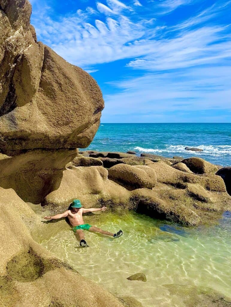 A child sitting in a clear rock pool between granite boulders at El Tule Beach