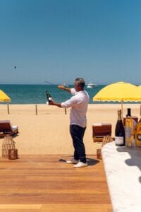 A server sabering a bottle of champagne on the deck at Delphine, with the beach, yellow umbrellas and yachts behind