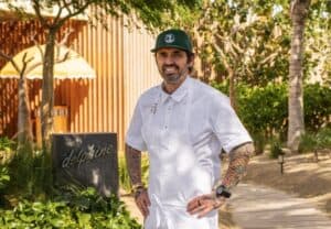 Chef Ludo Lefebvre in chef's whites and a cap beside the Delphine sign on the garden path at Costa Palmas