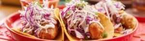 Crispy battered tacos topped with red cabbage slaw and crema on a red plate at El Barrio