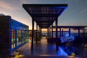 The covered walkway into El Barrio at dusk, with the restaurant's blue neon sign and the sea on the horizon
