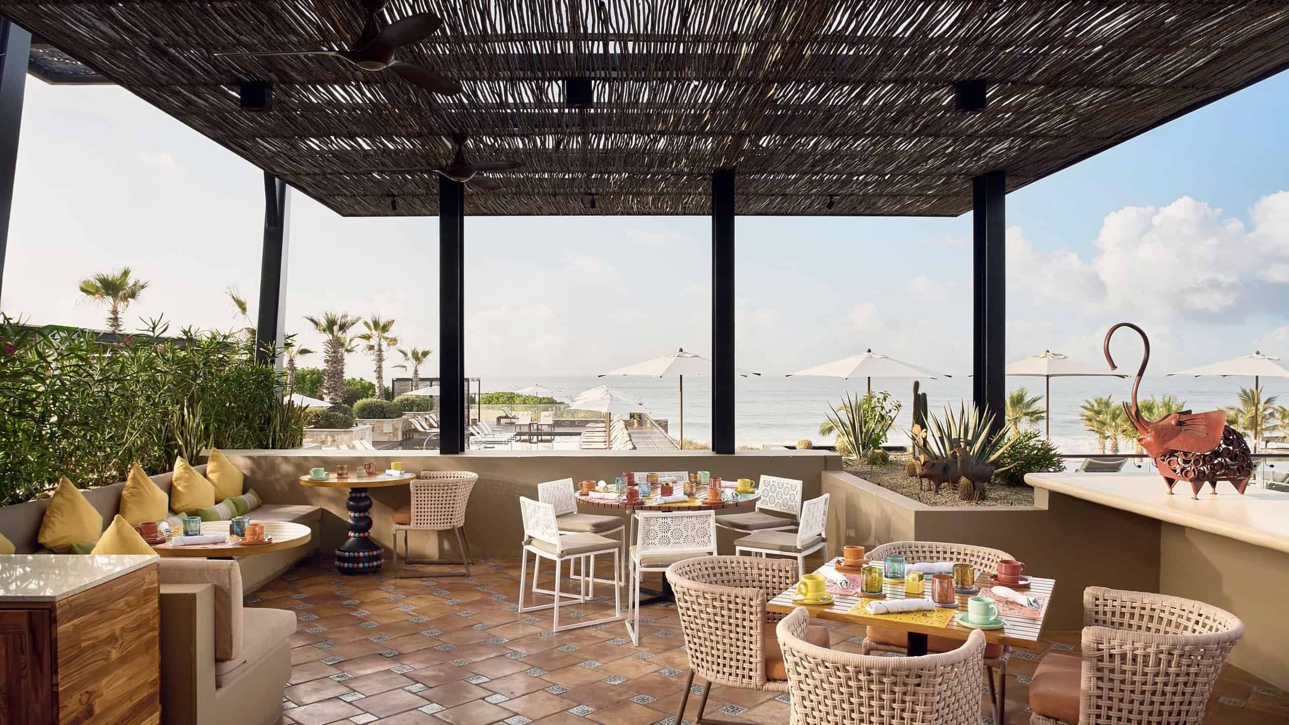 The covered terrace at El Barrio with woven chairs and colorful place settings, and the beach and the Sea of Cortez beyond, at Zadún in San José del Cabo