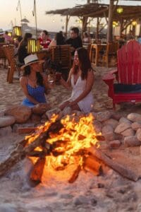 Two guests sitting by a fire pit on the sand at The Green Room, with the palapa and its lights behind them