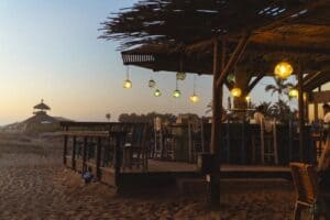 The palapa-roofed bar and wooden deck of The Green Room on the sand at dusk, with glass lanterns hanging from the roof