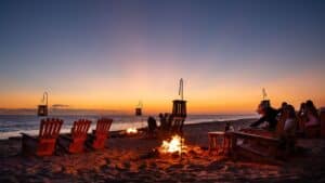 Guests in Adirondack chairs around fire pits on the sand at The Green Room as the sun sets over the Pacific