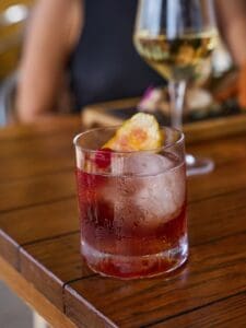 A red cocktail on ice with a dried citrus garnish on a wooden table at Jack's Steak House